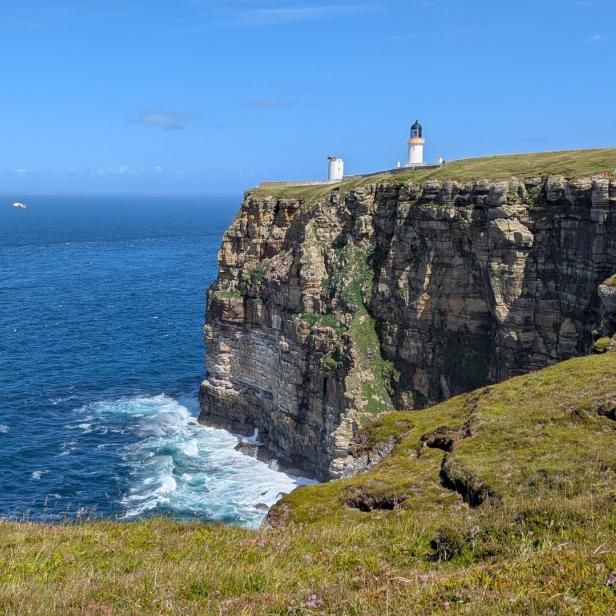 Steilküste am Meer mit Leuchtturm auf der Klippe und Wellenbrandung darunter.