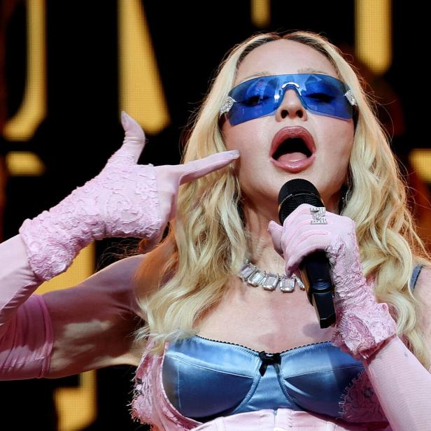 Madonna singt in ein Mikrofon, trägt eine blaue Sonnenbrille, ein blaues Korsett und rosafarbene Spitzenhandschuhe.