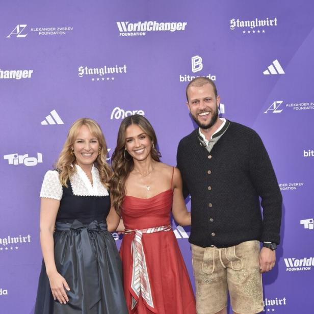 Zwei Frauen und ein Mann posieren vor einer violetten Wand mit Sponsorenlogos, wobei die Frauen in Dirndl und Kleid sowie der Mann in Lederhose auftreten.