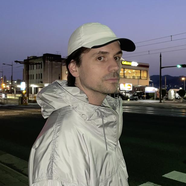 Mann in weißer Kappe und heller Jacke steht an einer nächtlichen Kreuzung mit Ampeln und Bergen im Hintergrund.