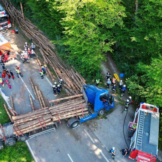 Luftaufnahme eines Unfalls mit umgestürztem Holztransporter, Feuerwehrleuten und mehreren Einsatzfahrzeugen auf einer Waldstraße.