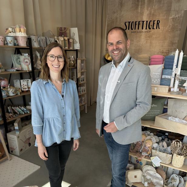 Zwei lächelnde Personen stehen in einem Baby- und Kindershop zwischen Regalen mit Geschenkartikeln und Deko.
