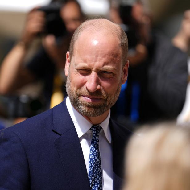 Prinz William trägt einen dunkelblauen Anzug mit Krawatte und blickt vor einer Menschenmenge mit Kameras zur Seite.