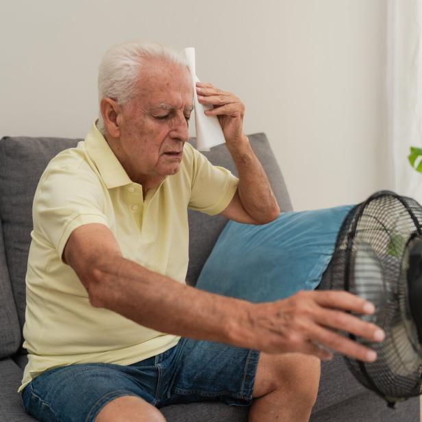 Senior man suffering from summer heat wiping sweat with napkin and holding a electric fan at home