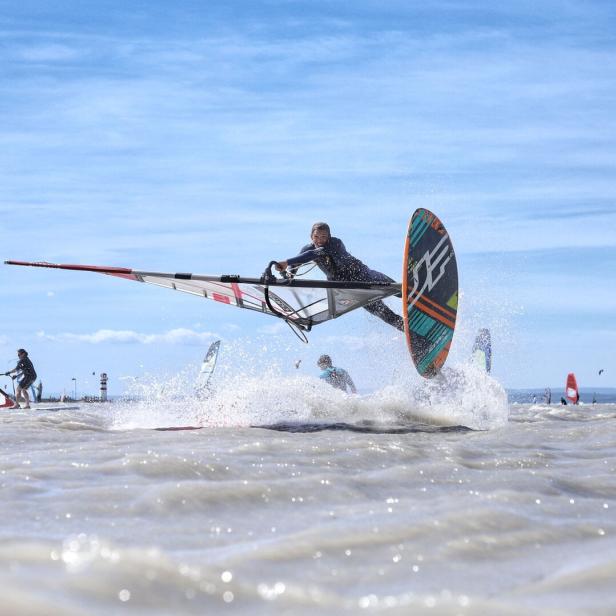 Windsurfer springt über eine Welle im Meer, umgeben von anderen Surfern.