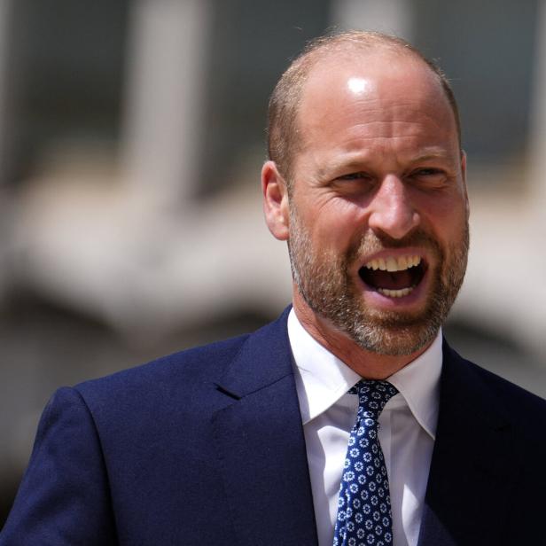 Prinz William trägt einen dunkelblauen Anzug mit weißem Hemd und gemusterter Krawatte und lacht mit offenem Mund.