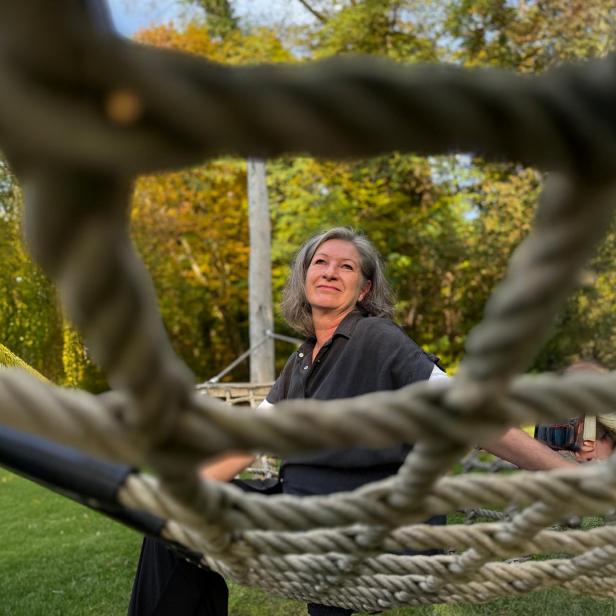 Frau sitzt auf einem Kletternetz im Freien und blickt lächelnd in die Ferne.