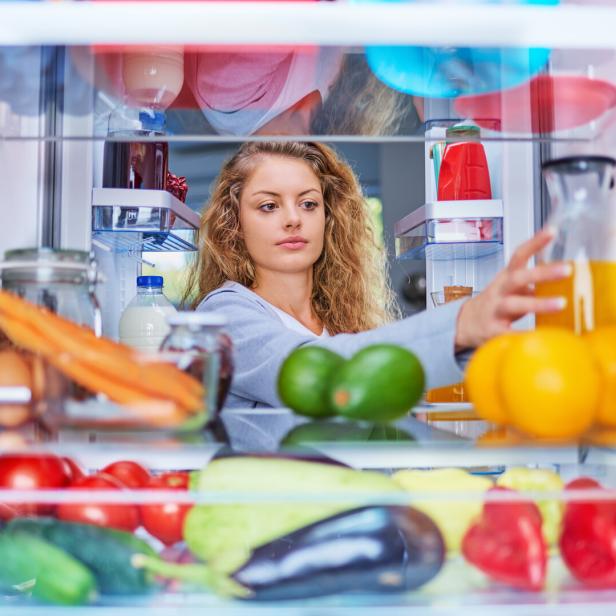 Eine junge Frau mit lockigem Haar greift aus der Perspektive des Kühlschrankinneren nach einem Glas Saft zwischen frischem Obst und Gemüse.