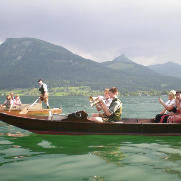 Mehrere Menschen in Tracht fahren und musizieren in Holzbooten auf einem See vor Bergen.