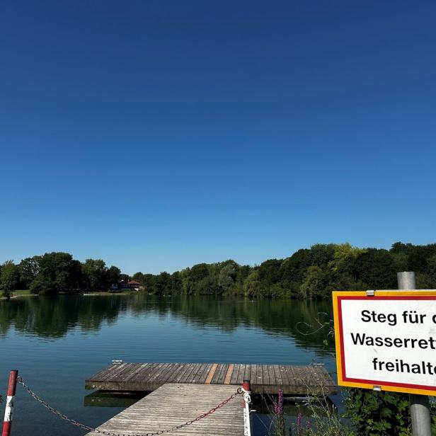 Ruhiger See mit Holzsteg und Schild „Steg für die Wasserrettung freihalten“.