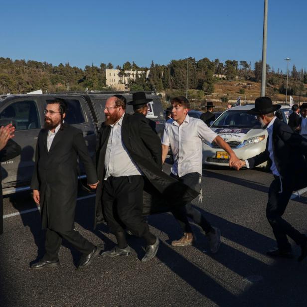 Eine Gruppe orthodoxer jüdischer Männer tanzt Händchen haltend auf einer Straße vor parkenden Autos.