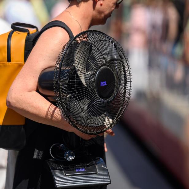 Symboldbild: Eine Person trägt einen gelben Rucksack und einen schwarzen Tischventilator der Marke SMC unter dem Arm.
