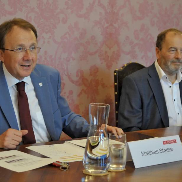 Zwei Männer im Anzug sitzen an einem Konferenztisch mit Wasserkrügen und Namensschild.