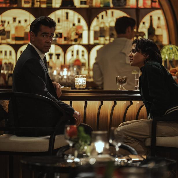 Colin Farrell im Anzug und eine Frau sitzen sich an einer beleuchteten Bar gegenüber und unterhalten sich.