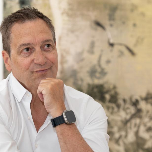 Dieter Nuhr in einem weißen Hemd stützt sein Kinn auf die Hand und blickt zur Seite vor einem abstrakten, hellen Hintergrund.