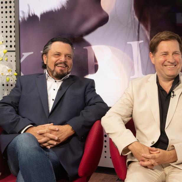 Werner Auer und Erich Furrer im Gespräch sitzen lachend in roten Sesseln, der eine in dunklem, der andere in hellem Anzug.