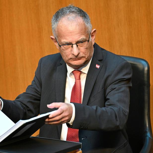 Markus Marterbauer liest in einer Mappe an einem Tisch vor braunem Hintergrund.