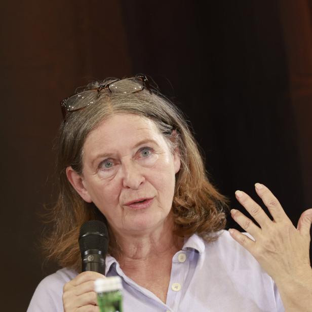 Eine Frau mit Brille auf dem Kopf spricht in ein Mikrofon und gestikuliert mit der Hand vor einem dunklen Hintergrund.
