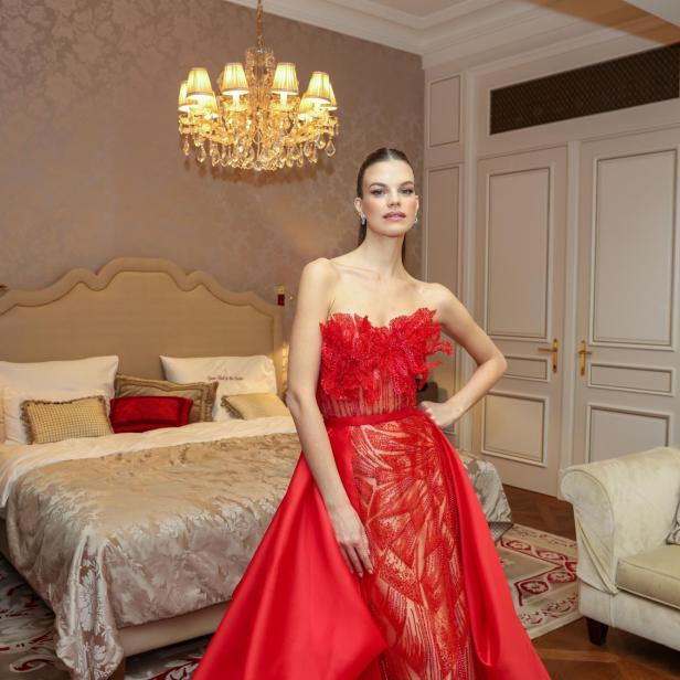 Eine Frau in einem eleganten, roten Abendkleid posiert in einem luxuriös eingerichteten Hotelzimmer vor einem Doppelbett.