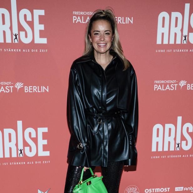 Lächelnde Frau in schwarzem Leder-Outfit mit grüner Handtasche vor pinker ARISE-Wand in Berlin.