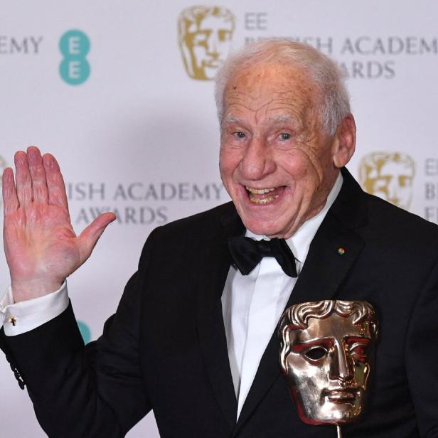 Mel Brooks lächelt fröhlich und winkt vor einer Kulisse der EE British Academy Film Awards mit einer BAFTA-Trophäe in der Hand.