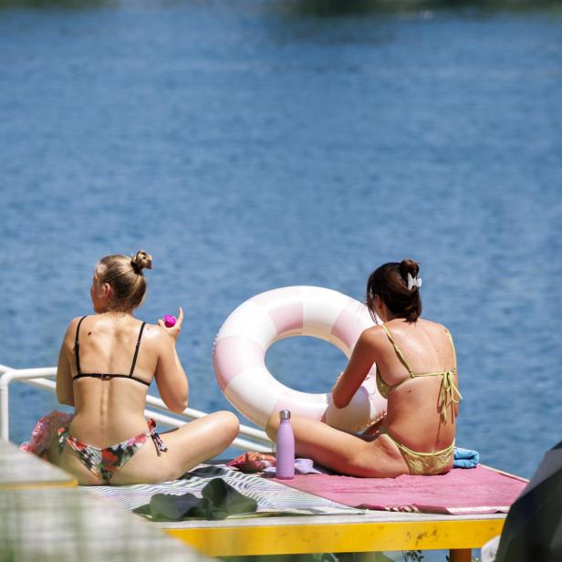 Zwei Frauen in Bikinis sitzen auf einem Steg am Wasser und halten einen rosa-weißen Schwimmreifen.