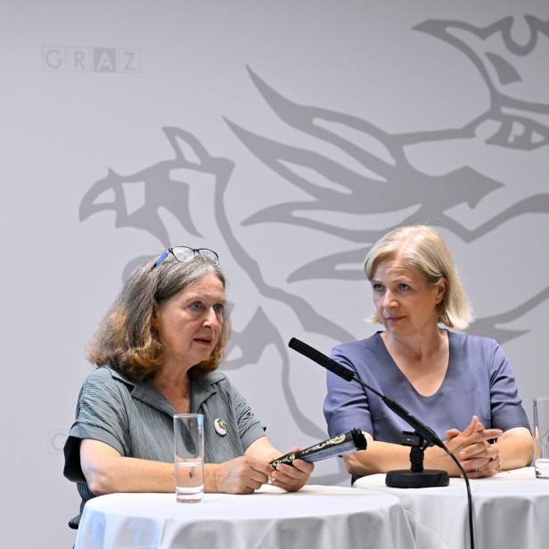 Zwei Frauen sitzen vor einem Hintergrund mit dem Grazer Stadtwappen an Stehtischen bei einer Pressekonferenz.