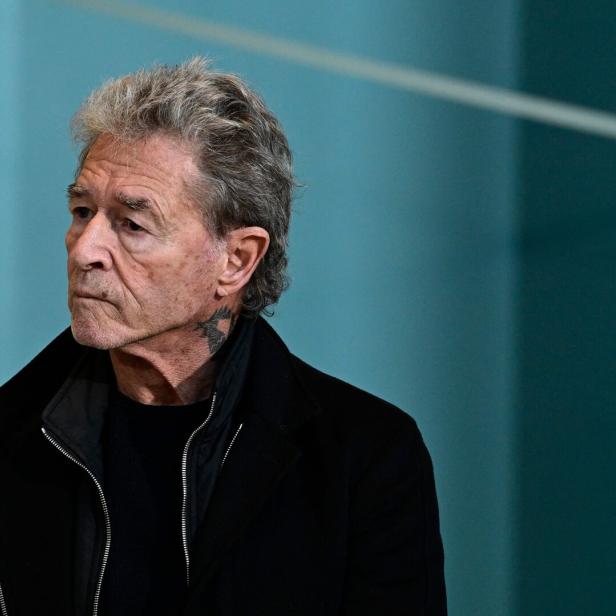 Peter Maffay mit grauem Haar und einem Tattoo am Hals vor einem türkisfarbenen Hintergrund.
