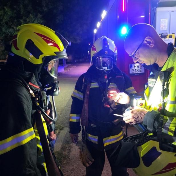 Drei Feuerwehrleute bei einer nächtlichen Einsatzbesprechung neben einem Fahrzeug mit Blaulicht.