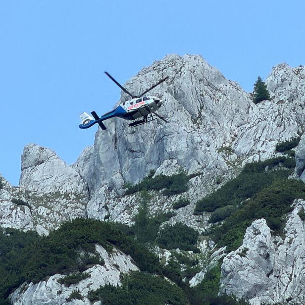 Hubschrauber fliegt über einer felsigen Berglandschaft mit wenigen Bäumen.