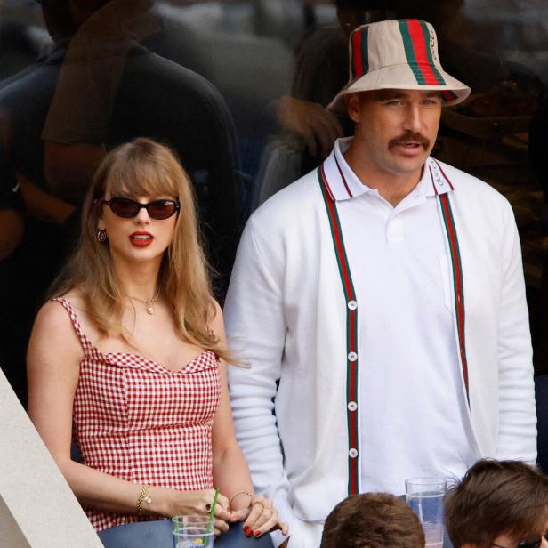 Taylor Swift und Travis Kelce sitzen nebeneinander auf einer Tribüne, wobei sie ein rot-weiß kariertes Kleid und er einen Gucci-Hut trägt.