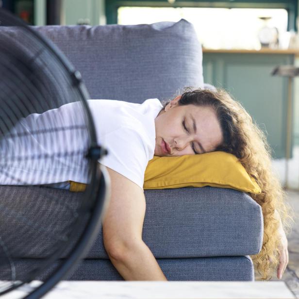 Eine Frau schläft erschöpft auf einer Couch, ihr Kopf liegt auf einem gelben Kissen, davor ein laufender Ventilator.