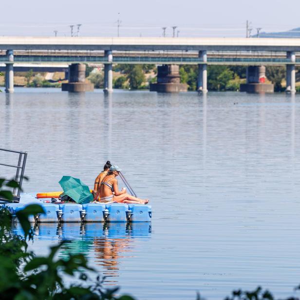 Zwei Frauen sitzen in Badekleidung auf einem blauen Schwimmsteg im Wasser vor einer großen Brücke.
