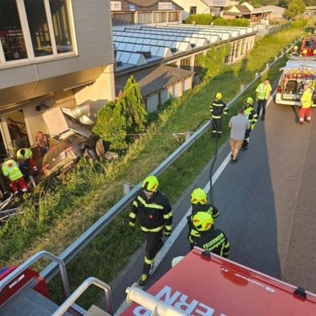 Feuerwehrleute und Rettungskräfte bergen ein Auto an einem Hang neben einem Gebäude; mehrere Einsatzfahrzeuge stehen auf der Straße.