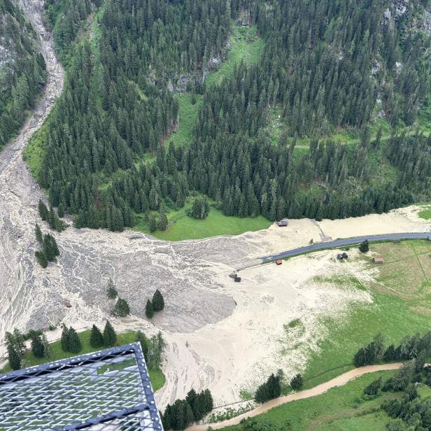 Eine Mure hat sich aus einem bewaldeten Berghang gelöst und eine Schlammlawine über eine Wiese und eine Straße in ein Tal gewälzt.