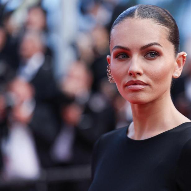Thylane Blondeau trägt ein schwarzes Kleid und blickt mit streng zurückgekämmtem Haar vor einem unscharfen Hintergrund in die Kamera.