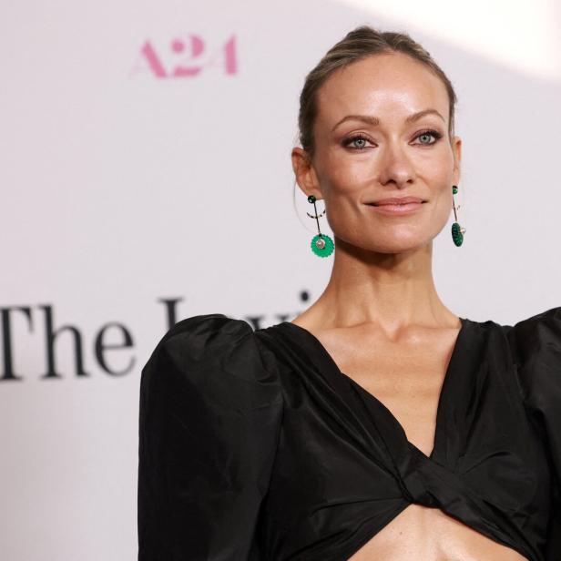 Olivia Wilde trägt ein schwarzes Oberteil und grüne Ohrringe vor einem A24-Hintergrund.