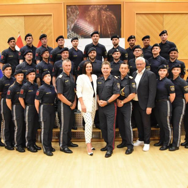 Polizeigruppe in Uniform posiert mit zwei Zivilpersonen in einem Saal vor EU- und Österreich-Flaggen.