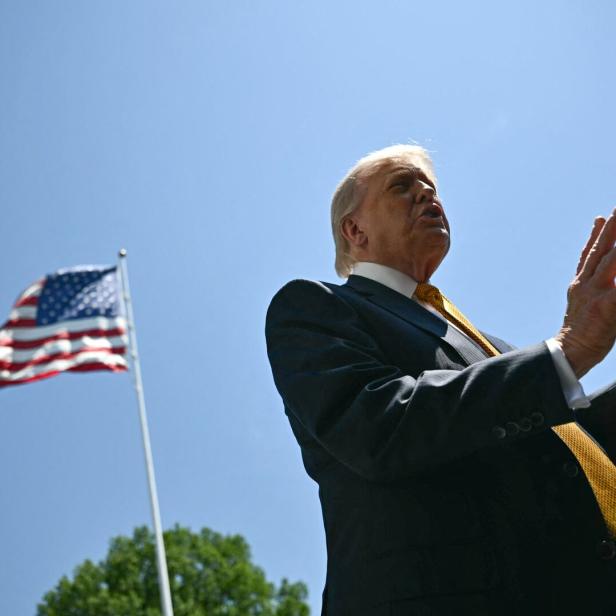 Donald Trump spricht vor einer wehenden US-Flagge unter blauem Himmel.
