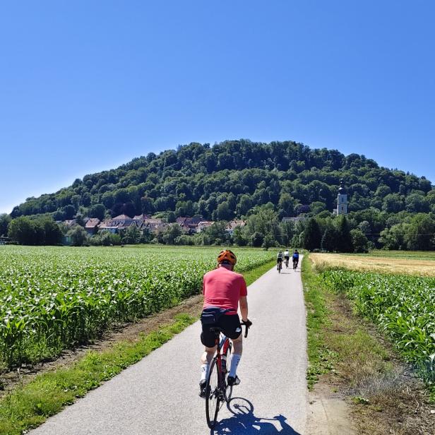Eine Gruppe von Radfahrern fährt auf einem asphaltierten Weg zwischen Feldern auf ein Dorf mit bewaldetem Hügel zu.