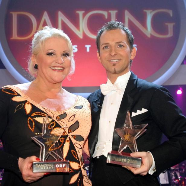 Marika Lichter und ihr Tanzpartner präsentieren stolz ihre Trophäen vor dem Logo der ORF-Show Dancing Stars.
