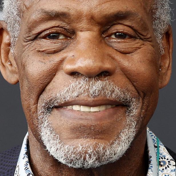 Nahaufnahme des lächelnden Schauspielers Danny Glover mit grauem Haar und Bart vor einem dunklen Hintergrund.