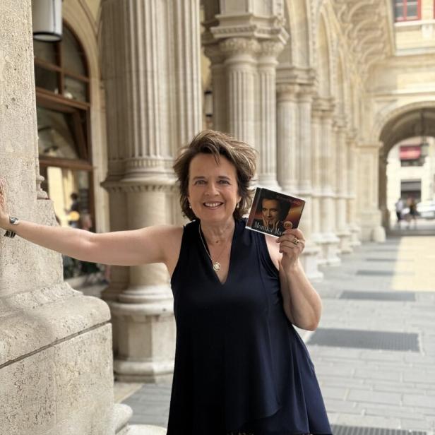 Frau im schwarzen Top und Leopardenhose steht in einer säulengesäumten Passage und hält eine CD hoch.