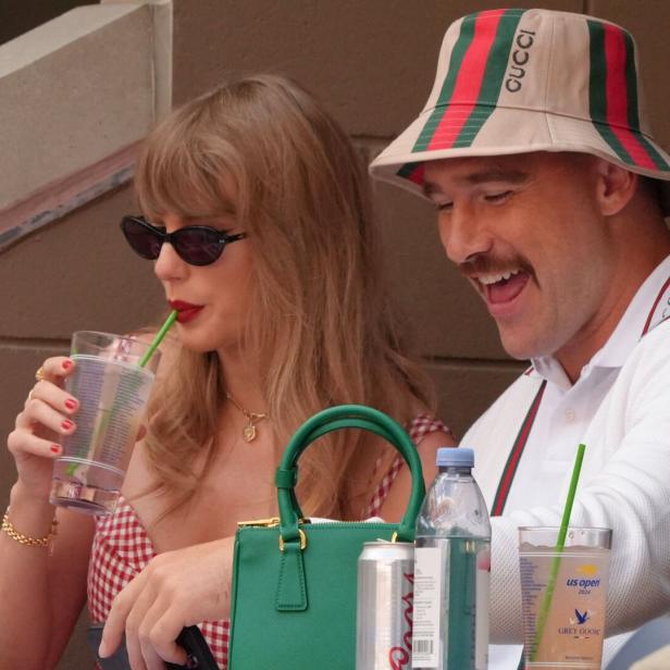 Taylor Swift und Travis Kelce sitzen nebeneinander bei den US Open, wobei Swift ein Getränk trinkt und Kelce lachend einen Gucci-Hut trägt.