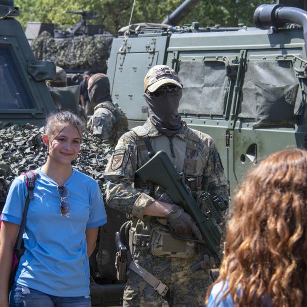 Ein bewaffneter Soldat in Tarnuniform steht vor Militärfahrzeugen, daneben ein lächelndes Mädchen in blauem Shirt.