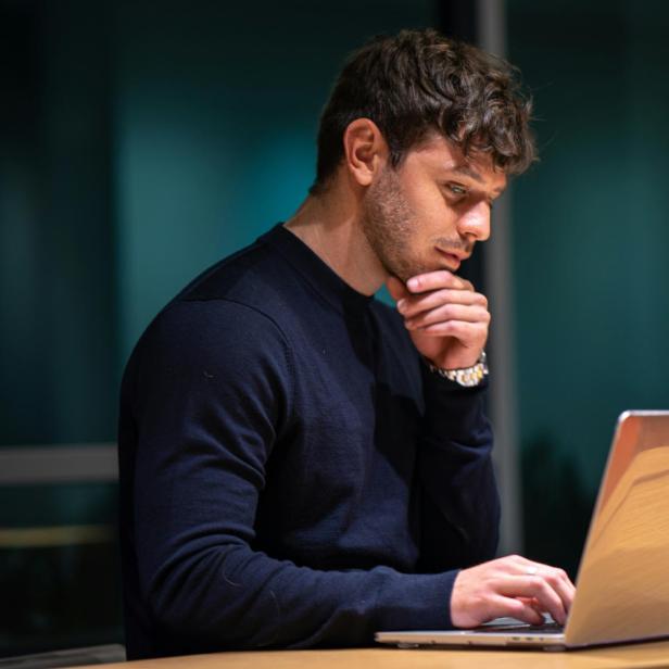 Mann mit dunklem Pullover sitzt nachdenklich vor einem Laptop und stützt das Kinn mit der Hand.
