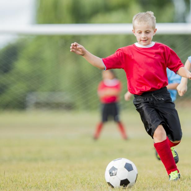 Mehrere Kinder spielen Fußball, ein Junge in Rot läuft zum Ball.