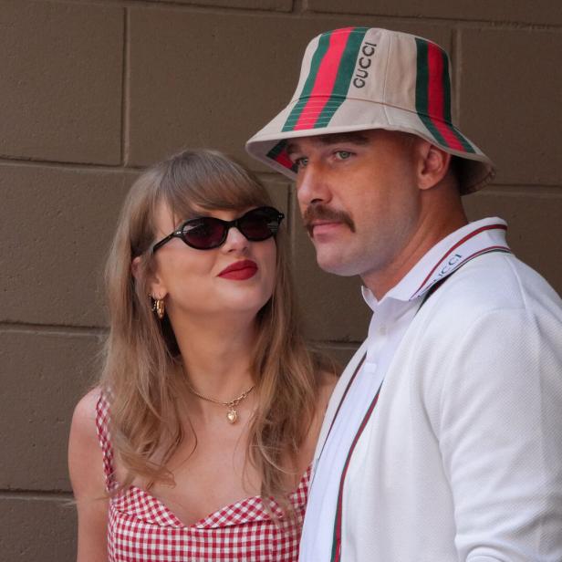 Taylor Swift mit Sonnenbrille blickt zu Travis Kelce, der einen Gucci-Fischerhut trägt, vor einer braunen Ziegelwand.