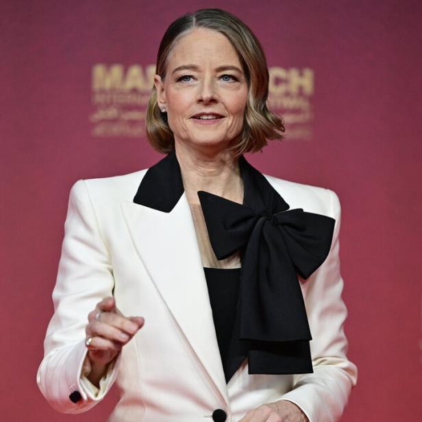 Jodie Foster trägt einen weißen Blazer mit schwarzem Revers und einer großen schwarzen Schleife vor einem roten Hintergrund.