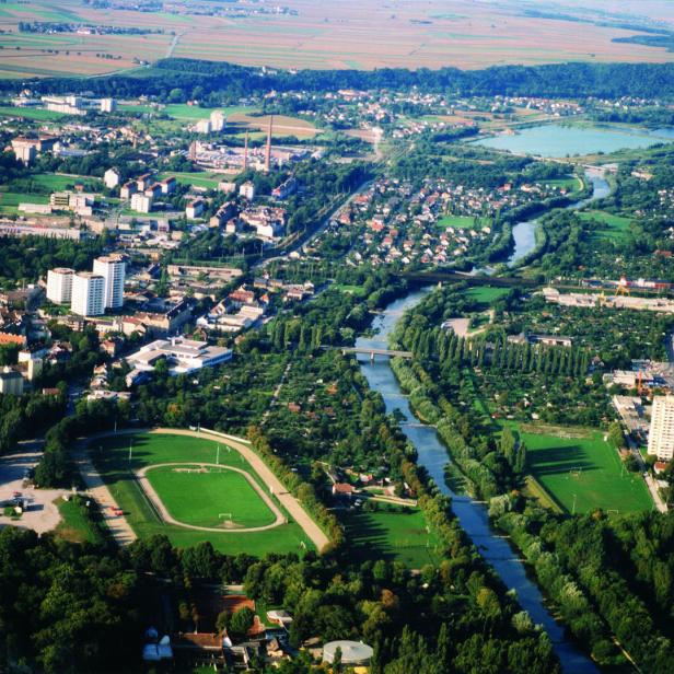 Luftaufnahme einer Stadt mit Fluss, vielen Grünflächen, Wohnhäusern und Feldern am Rand.